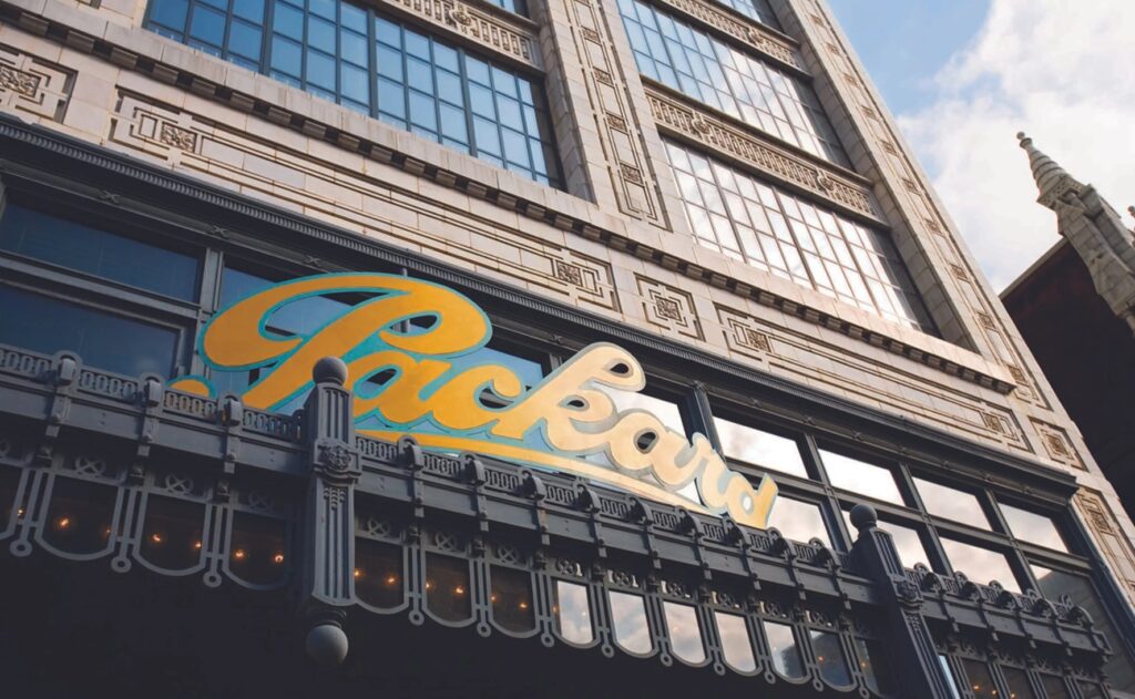 The exterior of a historic building with large windows and an ornate facade features a prominent yellow "Packard" sign above the entrance.