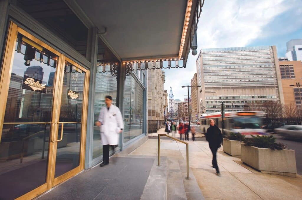 A person in a white coat exits a building onto a busy city sidewalk with blurred pedestrians and a bus passing on the street.