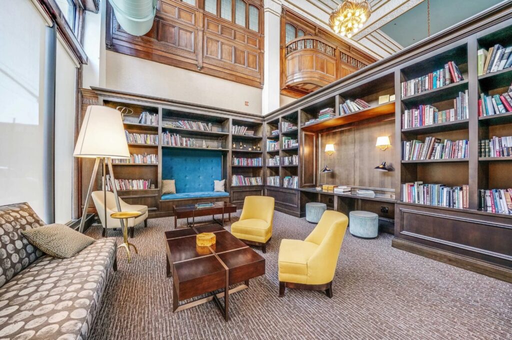 Spacious library lounge with bookshelves, patterned sofa, yellow armchairs, wood tables, and a reading nook under high ceilings with ornate trim and large windows.