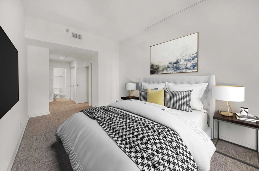 A modern bedroom with a neatly made bed, two nightstands with lamps, a large wall-mounted TV, and a door leading to a white bathroom.
