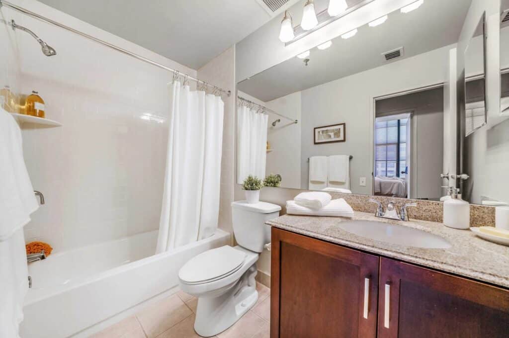 Modern bathroom with a shower-tub combo, white curtain, toilet, brown vanity with granite countertop, mirror, and towels neatly arranged.