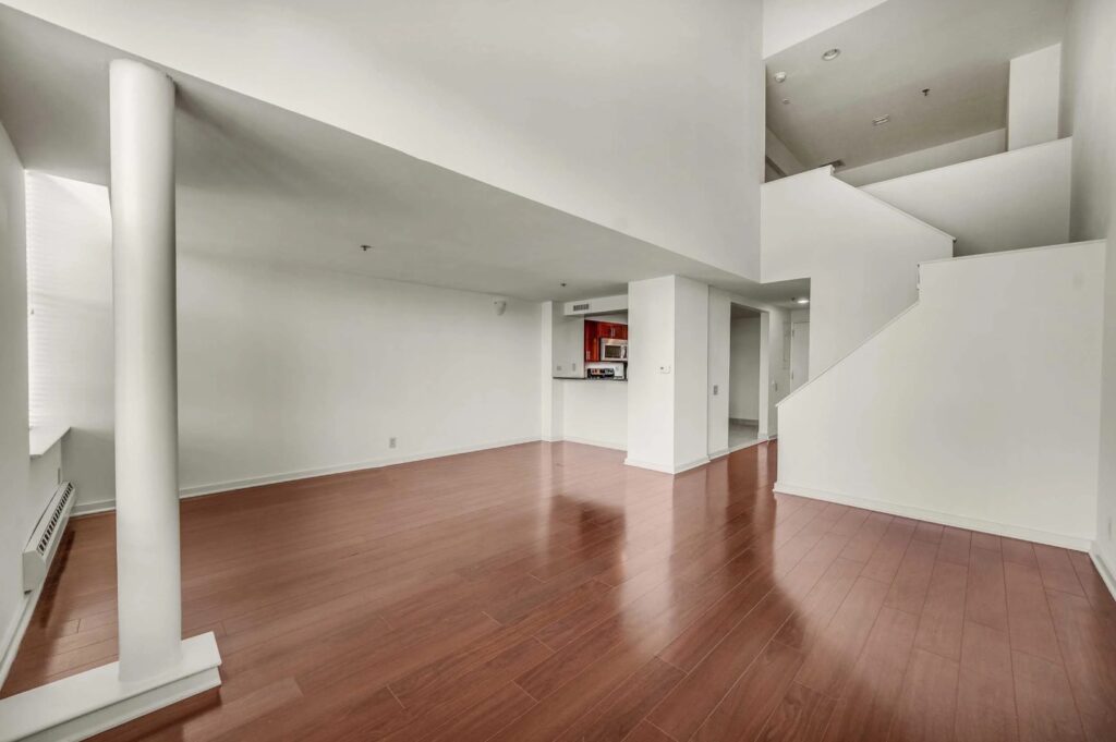 Spacious, empty room with polished wood flooring, white walls, a staircase, and an open view into a modern kitchen area with red cabinets.