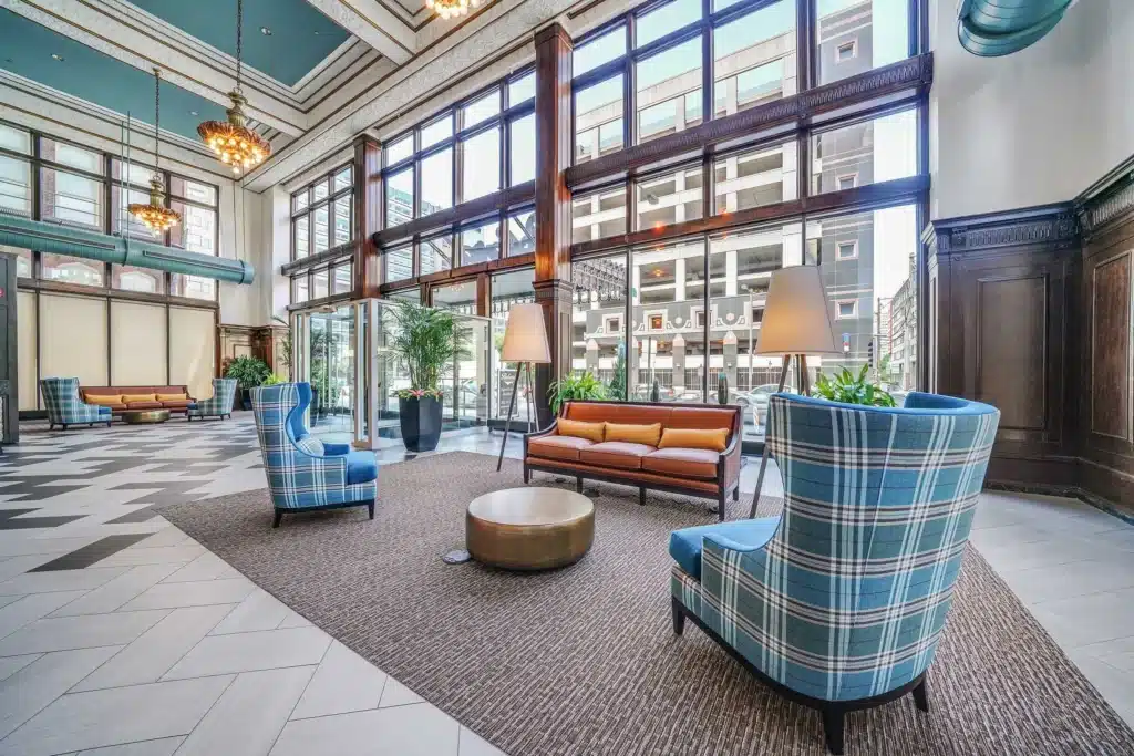 Spacious, modern hotel lobby with large windows, plaid armchairs, a brown sofa, indoor plants, pendant lights, and patterned tile flooring.