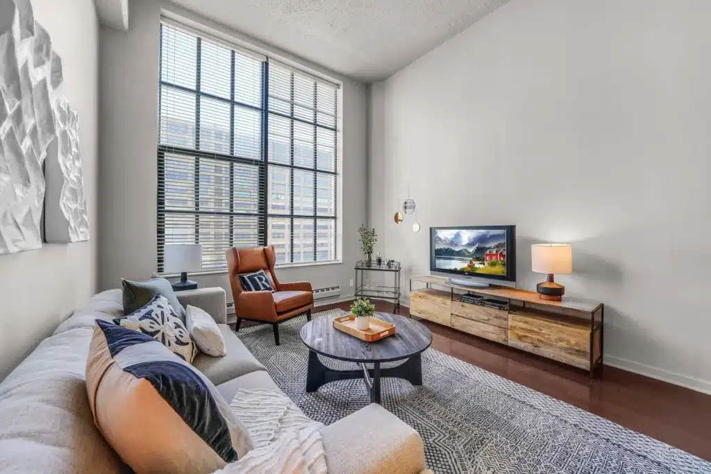 Modern living room with a large window, beige sofa, orange armchair, round coffee table, TV on a wooden stand, and decorative accents.