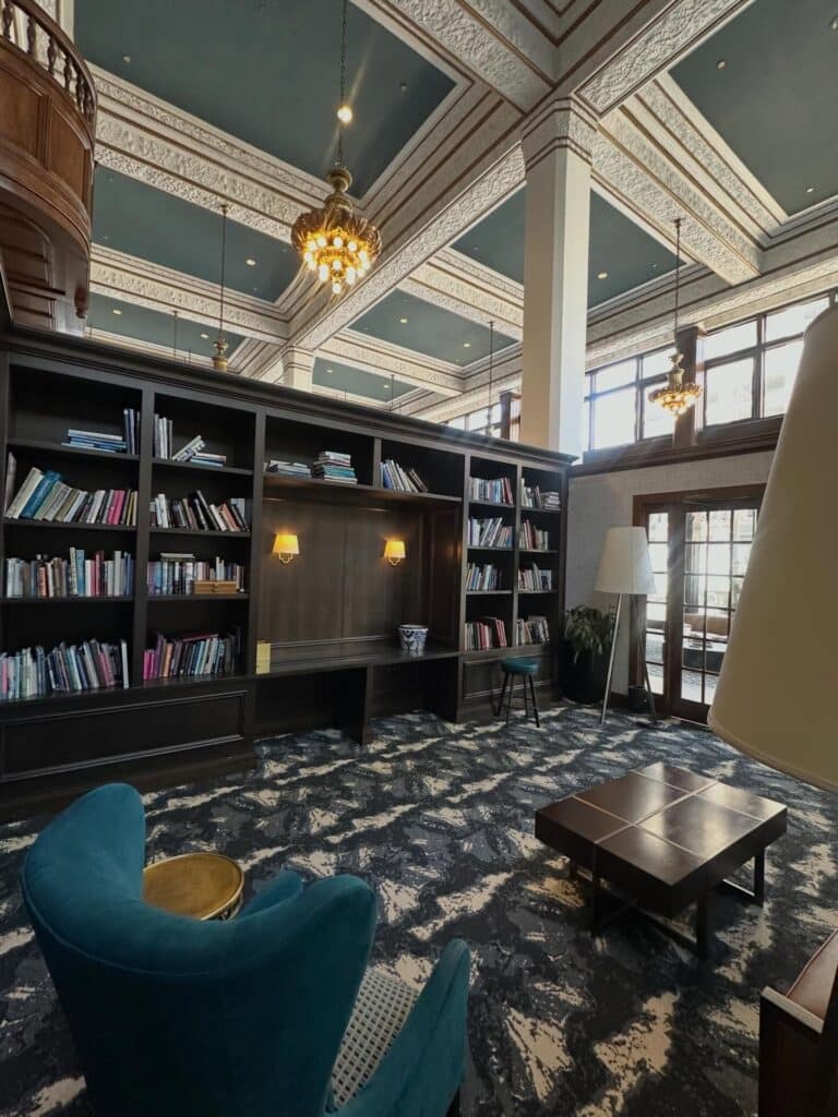 A spacious library room with tall ceilings, chandeliers, large windows, bookshelves, a teal armchair, modern furniture, and patterned carpet.